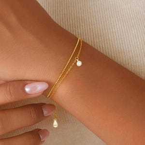 以下が含まれることがあります： 2本の細いチェーンが付いた繊細なゴールドブレスレット。1本のチェーンは点線で、もう1本はシンプルなリンクチェーンです。小さな丸い透明な宝石がリンクチェーンからぶら下がっています。涙型のチャームが留め金からぶら下がっています。