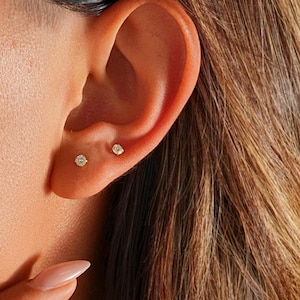 May include: Close-up of an ear with two small, gold stud earrings. The earrings have a single, clear gemstone. The ear is set against a background of brown hair and skin.