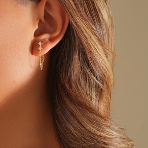 以下が含まれることがあります： 星型のデザインと繊細なチェーンが付いたゴールドのイヤリングのクローズアップ。イヤリングには、小さな輝く石が飾られています。イヤリングは、茶色の髪の人の耳からぶら下がっています。