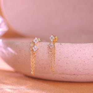 Puede incluir: Un par de pendientes dorados con diseño floral. Cada pendiente presenta una pequeña flor de cuatro pétalos con piedras claras y brillantes. Delicadas cadenas doradas cuelgan de las flores. Los pendientes se exhiben sobre una superficie rosa claro.