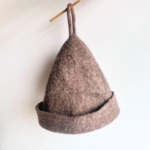 May include: A brown felt sauna hat with a conical shape and a rolled brim. The hat is suspended by a loop attached to a wooden dowel. The hat's texture appears soft and warm, suggesting it is made of wool.