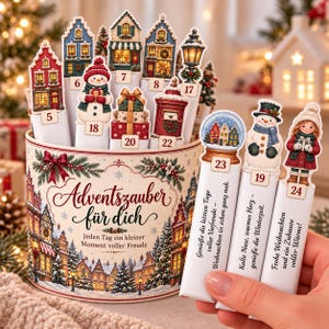 May include: A cylindrical advent calendar container with the text Adventszauber für dich. It features illustrations of winter houses and trees. Several numbered cardboard inserts with images of snowmen, houses, and gifts are tucked inside.