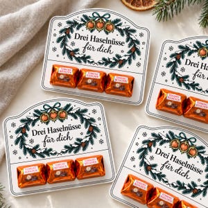 May include: Four rectangular white cards featuring a festive illustration of a pine wreath with three hazelnuts and the text "Drei Haselnüsse für dich". Each card holds three Ferrero Küsschen chocolates wrapped in shiny orange foil.