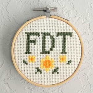May include: A cross-stitch embroidery hoop with the letters "FDT" in dark green thread. The design includes yellow and orange flowers with green accents. The hoop is made of light-colored wood and is set against a gray background.