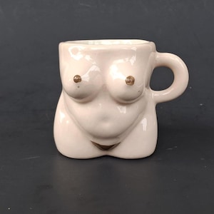 May include: A glossy, light-colored ceramic mug shaped like a human torso. The mug features a rounded handle on the right side, sculpted breasts with brown-painted nipples, a navel, and a small brown-painted triangle at the base of the torso.