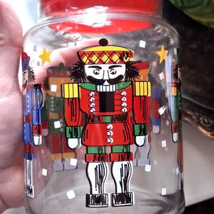 May include: Clear glass jar with a red lid, decorated with colorful nutcracker soldier illustrations and gold stars. The jar is a festive holiday decoration.