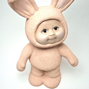 May include: A ceramic figurine of a baby in a bunny suit. The figure is primarily a light peach color, with a smiling face and large, expressive eyes. The bunny ears are upright and detailed.