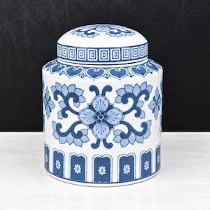 May include: A white ceramic ginger jar with a removable lid, featuring a blue floral pattern and a geometric border design. The cylindrical body is decorated with symmetrical blue leaf and flower motifs, while the base has a repeating arch pattern.
