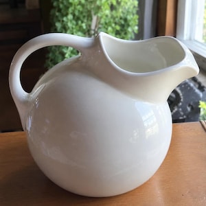 May include: A glossy white ceramic pitcher with a spherical, rounded body and a curved handle. The top features a wide, open mouth with a prominent, smooth pouring spout. The surface is solid white with a reflective finish and no patterns.
