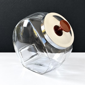 May include: A clear glass storage jar with a faceted, angular body. The jar features a circular, metallic silver lid topped with a small, round, dark brown knob handle. The container is positioned at an angle, showcasing its geometric glass design.