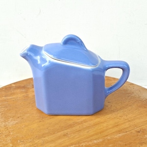 May include: A periwinkle blue ceramic teapot with a hexagonal body, a curved handle, a short spout, and a matching lid featuring a triangular handle. The teapot has a smooth, solid-colored glaze and a distinct geometric shape with flat sides.