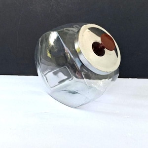 May include: A clear, vintage-style glass jar with a silver lid and a brown knob. The jar has a unique, geometric shape and is designed for storage. The lid has a silver rim and a brown center. The jar is empty.