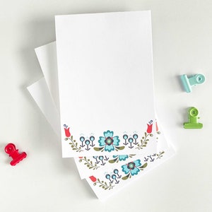 May include: A stack of white notepads with a floral border design. The border features red tulips, green leaves, and blue flowers. The notepads are on a white surface.