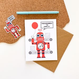 May include: A red robot-themed greeting card with the text "PROGRAMMED TO PARTY!" and a red balloon. A robot sticker with the text "BEEP BOOP" is on the left. A light blue pen rests on a corkboard.