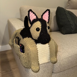 May include: A handmade crochet dog-shaped couch armrest organizer in tan, black, and white. The dog has black and pink ears, and a pocket on the side holds items like glasses and a remote. The dog is positioned on the arm of a beige couch.