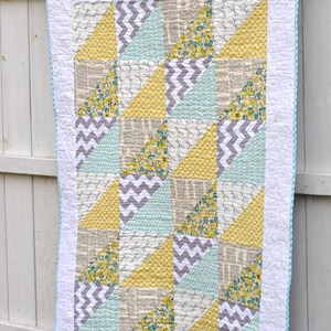 May include: A patchwork quilt with a white background and a blue and white checkered border. The quilt is made up of various geometric shapes in shades of yellow, gray, and blue. The quilt is hanging on a white fence.