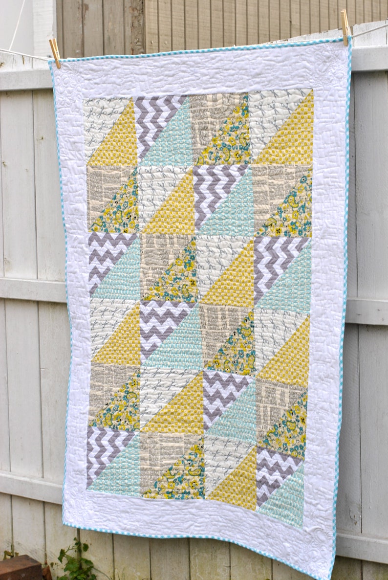 May include: A patchwork quilt with a white background and a blue and white checkered border. The quilt is made up of various geometric shapes in shades of yellow, gray, and blue. The quilt is hanging on a white fence.