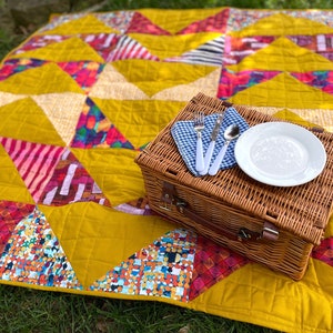May include: A yellow quilted picnic blanket with a colorful geometric pattern. A wicker picnic basket with a white plate, silverware, and a blue and white checkered napkin is on top of the blanket.