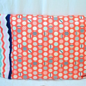 May include: A rectangular pillowcase with a coral background covered in white circles, each containing a different pattern. The left side features a vertical design of zigzags in blue, pink, and teal. A navy blue border separates the patterns.