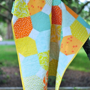 May include: A yellow, orange, and blue patchwork quilt with a white background. The quilt is made of hexagon-shaped fabric pieces and is hanging from a tree branch.