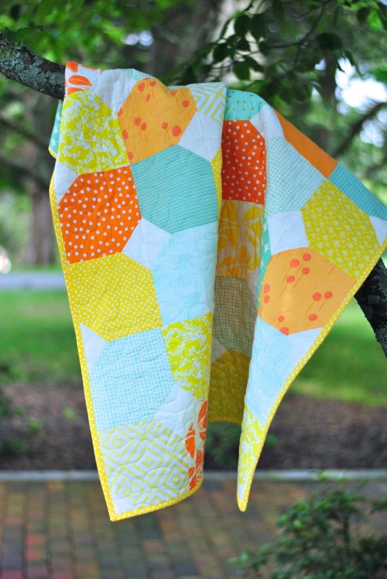 May include: A yellow, orange, and blue patchwork quilt with a white background. The quilt is made of hexagon-shaped fabric pieces and is hanging from a tree branch.