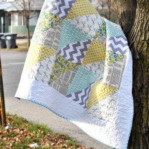 May include: A patchwork quilt with a white background and various colored squares. The squares are in shades of gray, yellow, and blue. The quilt is hanging from a tree branch.