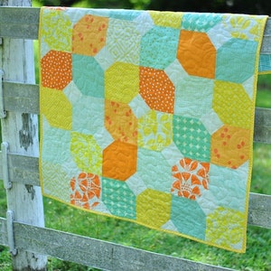May include: A colorful quilt with a geometric pattern in shades of yellow, orange, and turquoise. The quilt is hanging on a wooden fence.