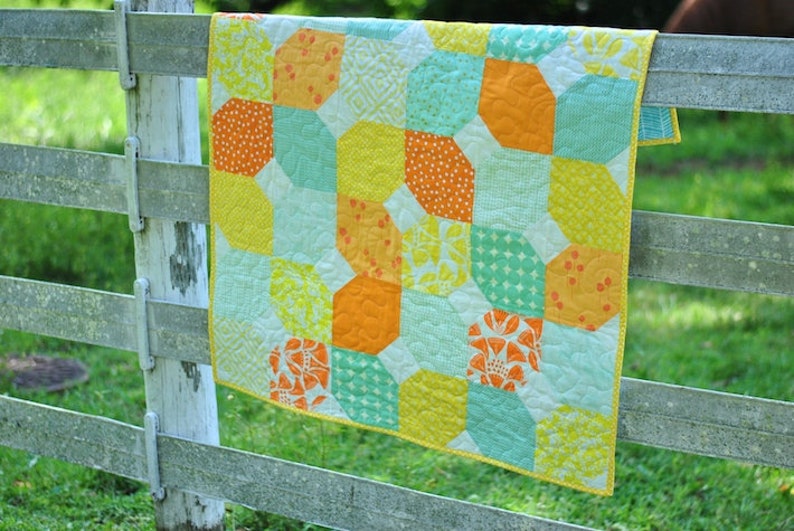 May include: A colorful quilt with a geometric pattern in shades of yellow, orange, and turquoise. The quilt is hanging on a wooden fence.