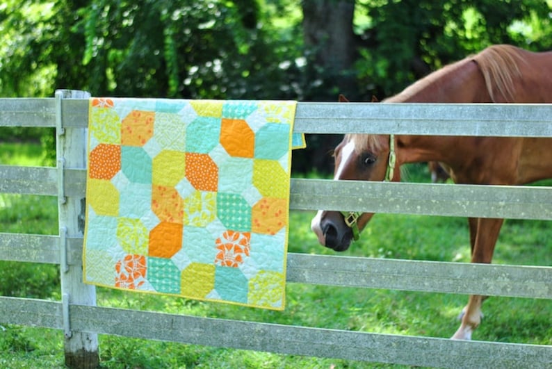 May include: A colorful patchwork quilt with yellow, orange, blue, and green squares hangs on a wooden fence. A brown horse with a white blaze on its face looks over the fence.
