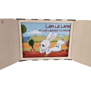 Può includere: Una scatola di legno aperta che mostra un libro per bambini. La copertina del libro presenta un coniglio bianco e il testo "LAPI LE LAPIN ET LES LÉGUMES DU JARDIN". L'illustrazione colorata mostra una scena di giardino.