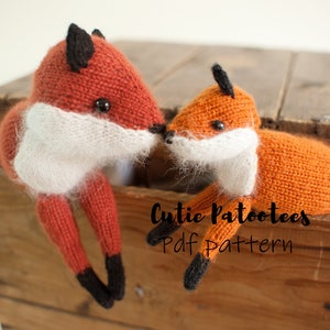 May include: Two knitted fox plushies, one larger than the other, are nestled in a wooden crate. The foxes are orange with white fur around their necks and black noses and eyes. The text "Cutie Patootees Pdf Pattern" is printed on the crate.