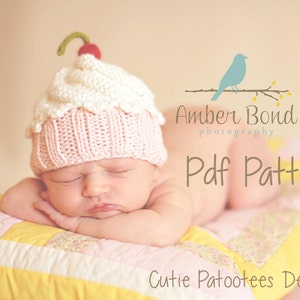May include: A newborn wearing a handmade knitted cupcake hat with a cherry on top. The hat is pink and white. The baby is lying on a patchwork quilt. The image includes the text "Pdf Pattern".