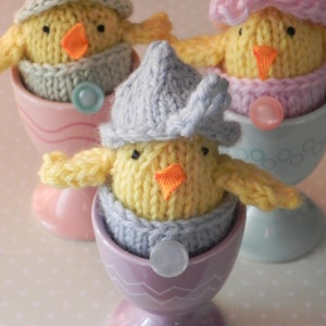 Könnte beinhalten: Drei gestrickte Küken in Eierbechern. Die Küken tragen Hüte und sind gelb mit orangefarbenen Schnäbeln. Die Eierbecher sind rosa, blau und lila.