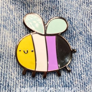 May include: A Non-binary pride bee enamel pin with a yellow head, white stripe, purple stripe, and black body. The bee has light green wings and a smiling face. The pin is on a blue denim background.