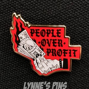 May include: A red and gold enamel pin with the words "PEOPLE OVER PROFIT". The pin depicts a hand holding burning money, with a skull on the notes. The pin is from "LYNNE'S PINS SHOP".