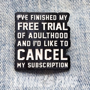 May include: A black and white rectangular pin with the text "I'VE FINISHED MY FREE TRIAL OF ADULTHOOD AND I'D LIKE TO CANCEL MY SUBSCRIPTION." The pin is set against a denim background.