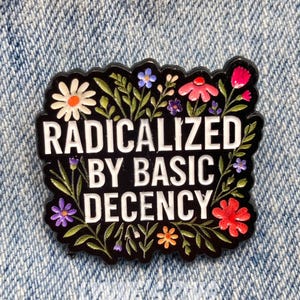 May include: A black enamel pin with the text "RADICALIZED BY BASIC DECENCY" in white. The pin is decorated with colorful flowers and green leaves. The pin is set against a blue denim background.