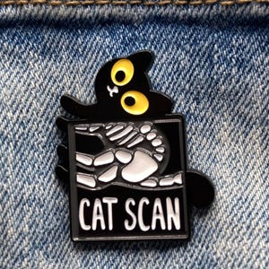 May include: A black enamel pin featuring a cartoon cat with yellow eyes and a skeleton inside a rectangle. The rectangle has the words "CAT SCAN" in white letters. The pin is on a denim background.