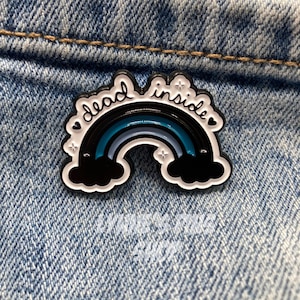 May include: A black, white, and blue enamel pin with the words "dead inside" arching over a rainbow design. The pin features black clouds at each end of the rainbow and is set against a denim background.