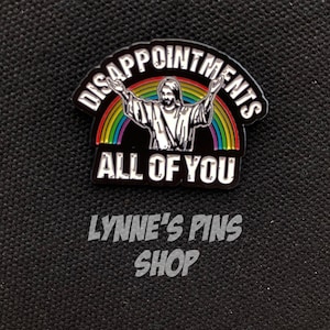 May include: A black and white enamel pin with a rainbow and a figure of Jesus with the text "Disappointments All Of You".