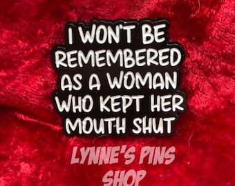 May include: Black enamel pin with white text that reads "I won't be remembered as a woman who kept her mouth shut". The pin is on a red velvet background. The text "LYNNE'S PINS SHOP" is also visible.