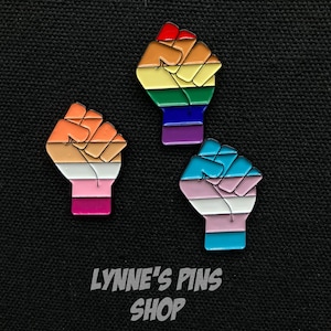 May include: Three enamel pins in the shape of a raised fist, each with a different LGBTQ+ pride flag design. One features a rainbow, another the lesbian flag, and the third the transgender flag. The text "LYNNE'S PINS SHOP" is at the bottom.