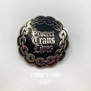 Peut inclure: Épinglette ronde argentée avec un centre noir. Le texte "Protect Trans Lives" est inscrit en gothique, entouré d'une chaîne. Le nom de la boutique "LYNNE'S PINS SHOP" est en bas.