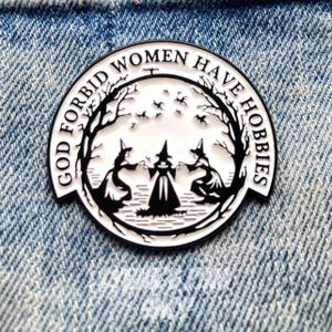 May include: A circular enamel pin, black and white, featuring the text "GOD FORBID WOMEN HAVE HOBBIES" around a scene of three witches and bats. The pin is set against a denim background.