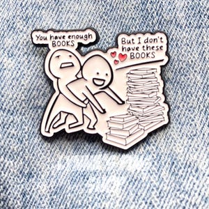 Peut inclure: Une broche en émail blanc avec des contours noirs représentant deux personnages de dessin animé et une pile de livres. Un personnage dit, You have enough BOOKS, et l'autre, But I don't have these BOOKS, avec trois petits cœurs rouges.
