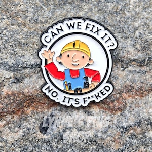 Peut inclure: Un pin's en émail circulaire représentant un personnage de dessin animé portant un casque de chantier jaune, une chemise rouge et une salopette bleue. Le texte CAN WE FIX IT? est arqué au-dessus et NO, IT'S F**KED en dessous du personnage.