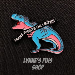 May include: A blue, pink, and white dinosaur enamel pin with the text "TRANS RIGHTS OR I BITES" on a black banner. The pin has a red mouth and is set against a black background. The text "LYNNE'S PINS SHOP" is at the bottom.