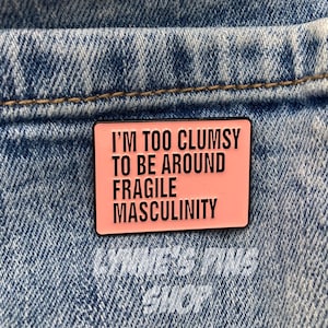 Peut inclure: Une épingle en émail rose avec une bordure noire et le texte "Je suis trop maladroit pour être autour de la masculinité fragile".