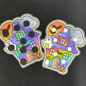 May include: Two die-cut stickers featuring a haunted house design with a rainbow gradient, ghosts, bats, and pumpkins. The left sticker has black scratch-off circles and text reading Save $50. The right sticker has white circles and text reading Roll & Save.