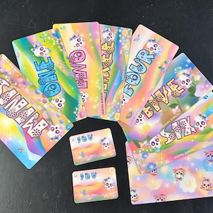 May include: A collection of colorful cards with a rainbow gradient design. The cards feature the words "SANMAN" and numbers "ONE" through "SIX" in a whimsical font, along with sugar skull illustrations and floral accents. Two smaller cards with "IOU" text are also present.
